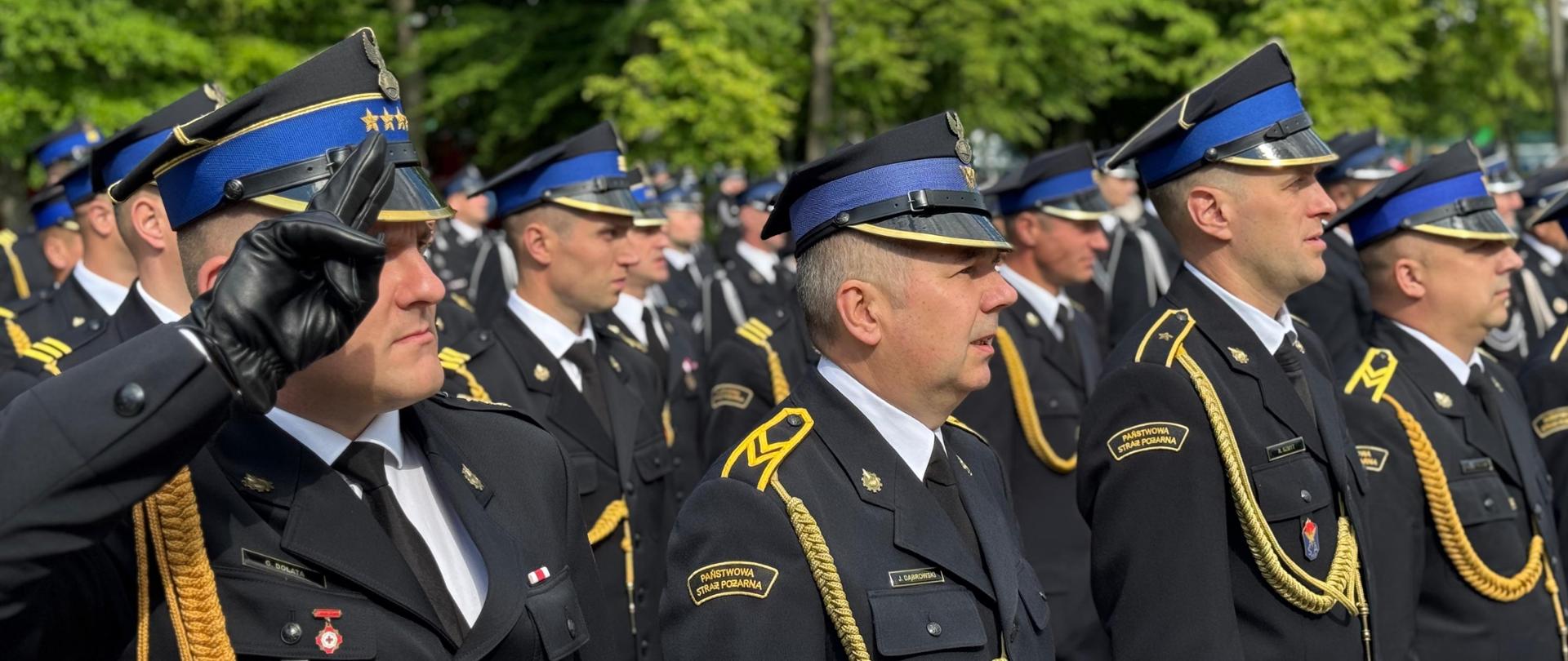 Na zdjęciu widoczni strażacy ubrani w mundury wyjściowe ze sznurem stojący podczas uroczystego apelu. Dowódca pododdziału oddaje honor pozostali strażacy stoją na baczność. 