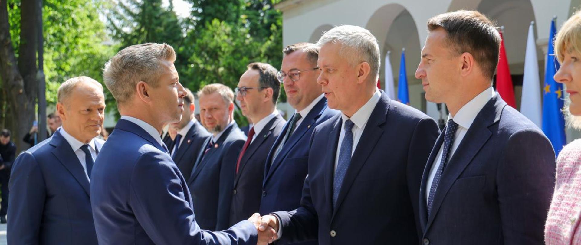 Na zdjęciu widać witających się ministra Tomasza Siemoniaka (po prawej) i premiera Węgier Pétera Magyara (po lewej). Za premierem Węgier stoi premier Donald Tusk. Po prawej i lewej stronie ministra Tomasza Siemoniaka stoją w szeregu inni ministrowie polskiego rządu. W tle widać budynek, drzewa i flagi.