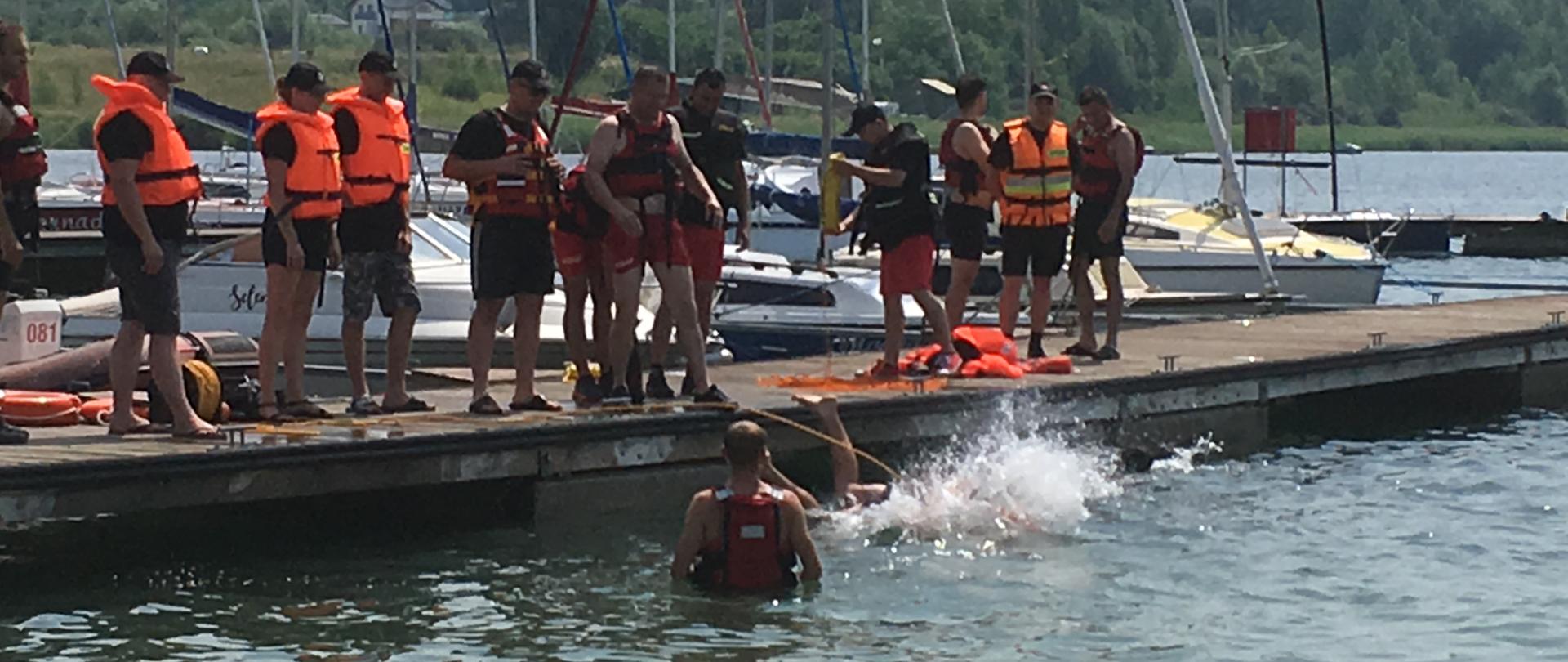 Pomost na jeziorze, na którym stoją ratownicy w kamizelkach ratowniczych. Jeden z mężczyzn wskoczył do wody. Przy pomoście widoczne są zacumowane jachty i łodzie. 