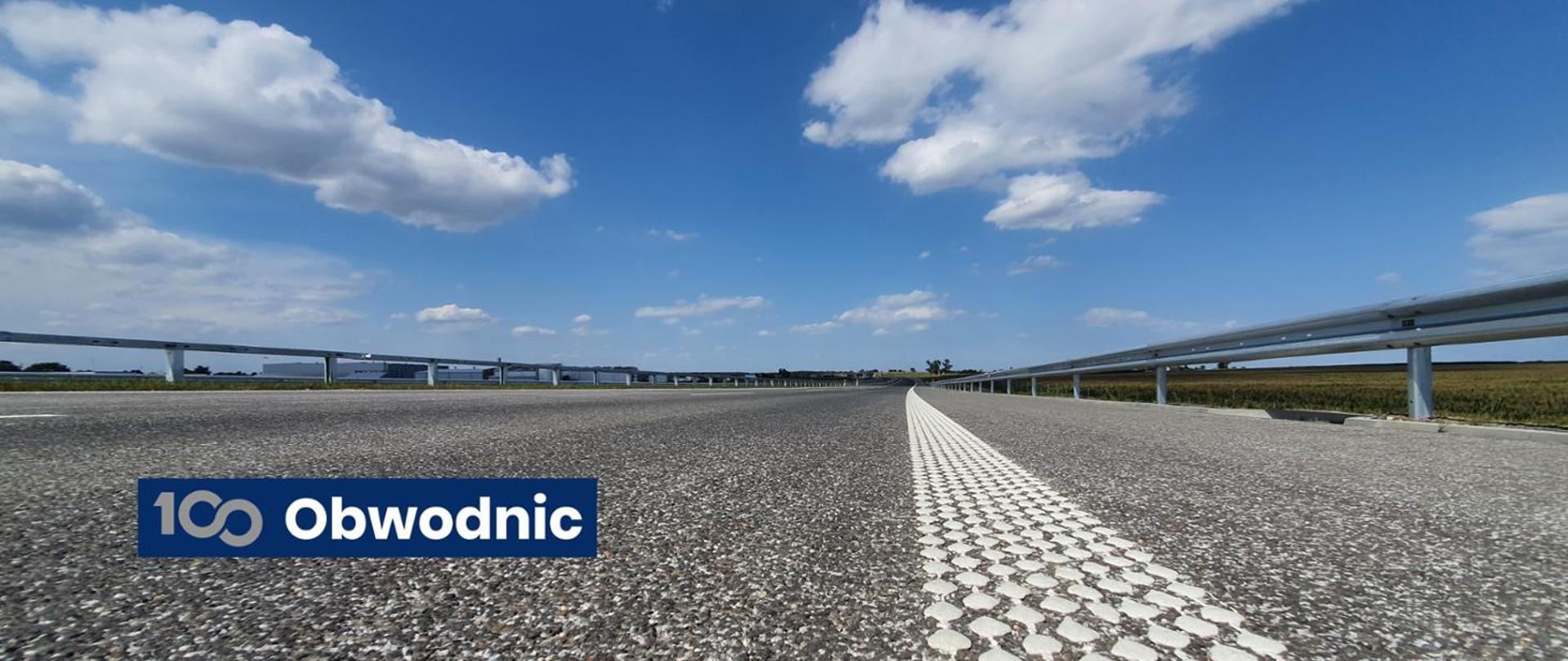 Widok z poziomu jezdni na nową obwodnicę w słoneczny dzień, z wyraźną białą linią przerywaną na środku asfaltu, metalowymi barierkami po obu stronach i niebieskim logo programu 100 Obwodnic w lewym dolnym rogu. W tle widać most i otwartą przestrzeń pod niebem z białymi chmurami.