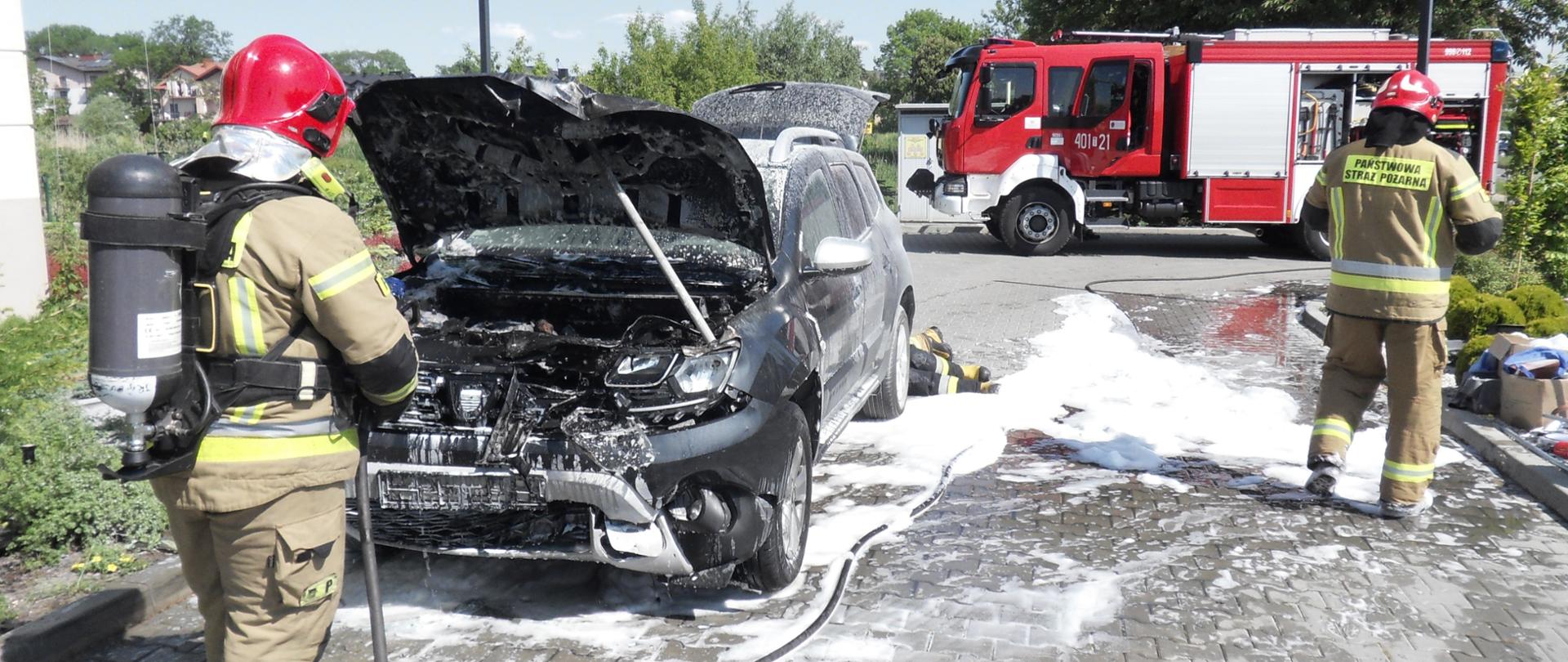 Po lewej stronie strażak PSP ubrany w ubranie ochronne koloru piaskowego z aparatem ochrony dróg oddechowych na plecach stojący tyłem i gaszący pożar komory silnika samochodu osobowego. Na kostce brukowej piana gaśnicza oraz po prawej stronie strażak PSP, w tle samochód pożarniczy od którego jest rozwinęty waż. 