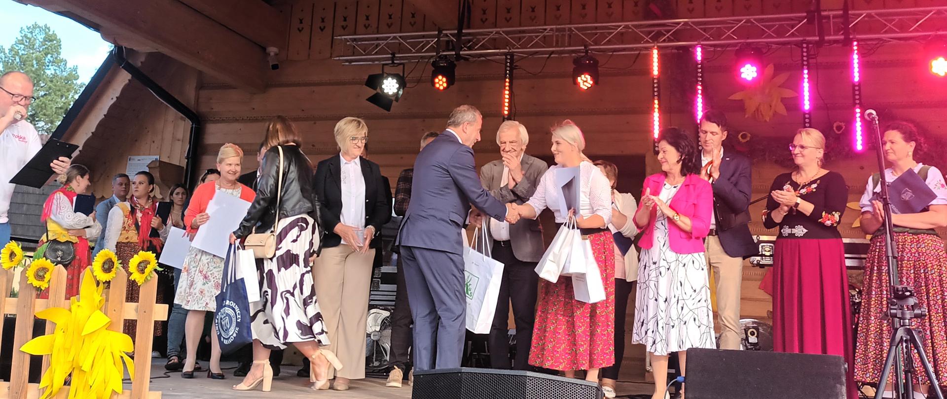 Na scenie mężczyzna stojący tyłem wręcza kobiecie nagrodę za udział w konkursie – w tle pozostali uczestnicy konkursu.