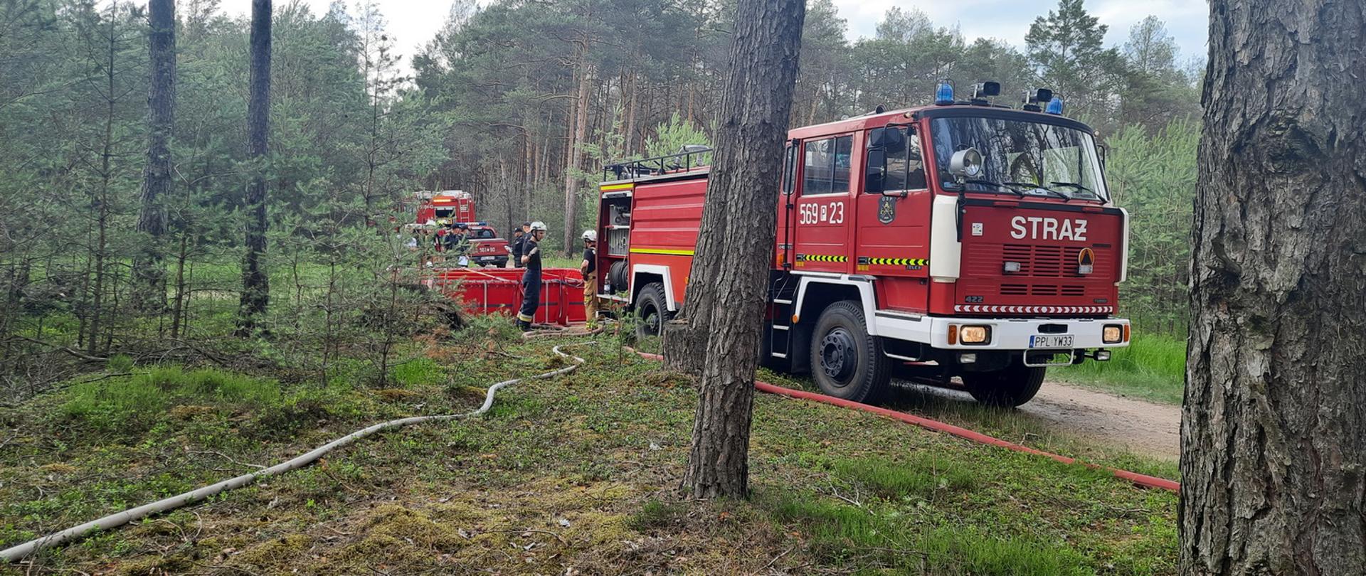 Wóz strażacki stoi na leśnej ścieżce, za nim widoczny brezentowy zbiornik na wodę i znajdujący się przy nim strażacy