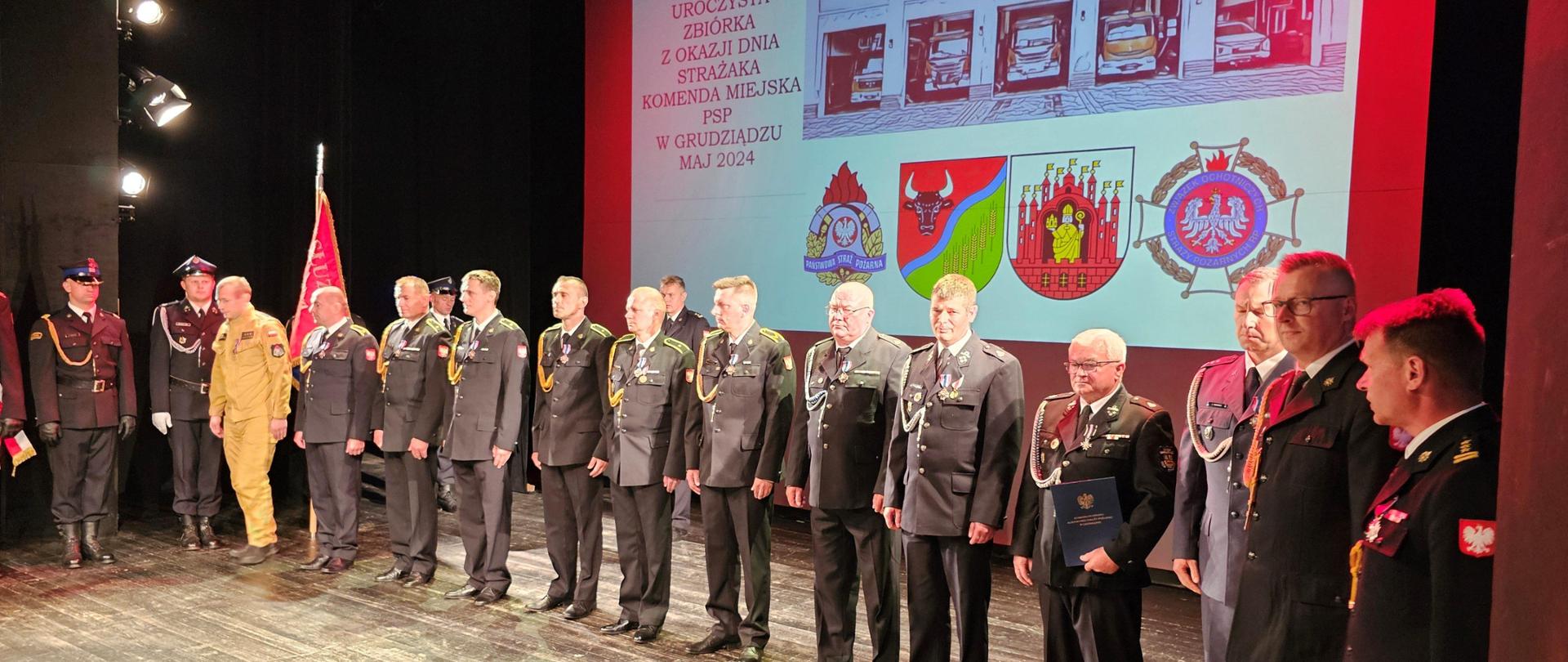 Strażacy stojący na scenie w rzędzie w galowych mundurach. W tle wyświetlana grafika dotycząca tej uroczystej zbiórki.