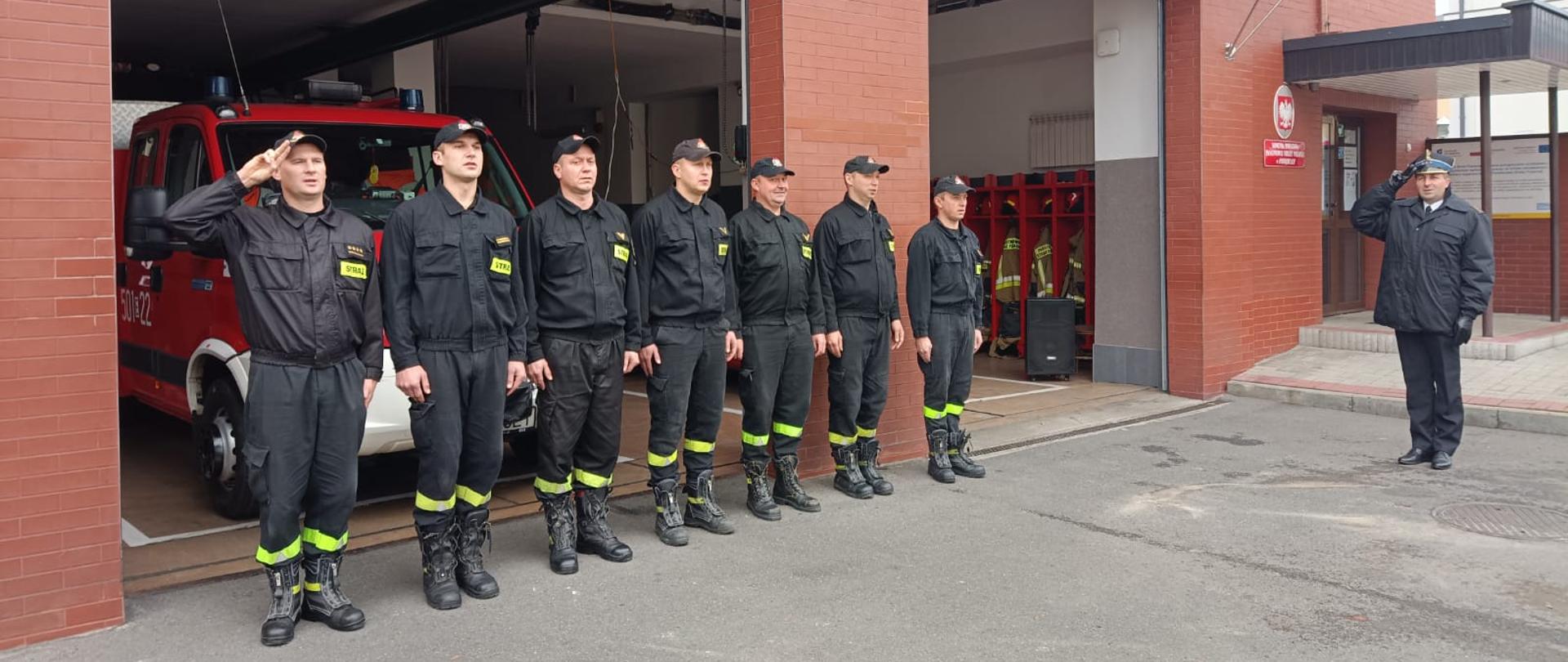 przed budynkiem komendy w jednym szeregu stoi 7 strażaków na baczność. Jeden z nich salutuje. Przed nimi stoi jeden funkcjonariusz również salutuje. Bramy wjazdowe do garaży są otwarte. Z tyłu widać czerwony samochód