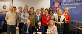 Grupowe zdjęcie uczestników spotkania w sali konferencyjnej. Kilkanaście osób stoi i klęczy w dwóch rzędach przed ekranem projekcyjnym. W tle widoczne są banery Wojewódzkiej Stacji Sanitarno‑Epidemiologicznej w Białymstoku.