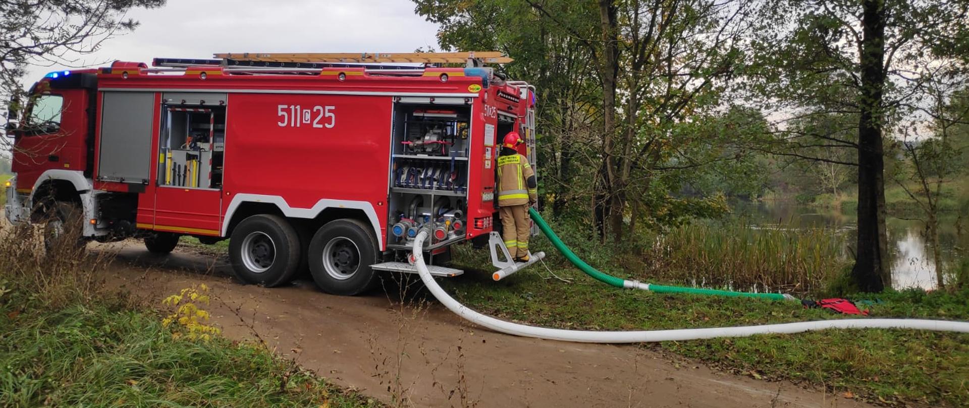 Samochód pożarniczy Volvo stoi na drodze gruntowej i ssie wodę z naturalnego zbiornika, następnie tłoczy ja do kolejnych, nie widocznych już na zdjęciu pojazdów pożarniczych.