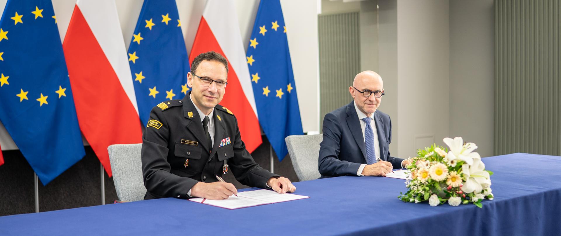 Komendant główny Państwowej Straży Pożarnej oraz Prezes Zarządu ORLEN podpisują dokumenty przy stole. W tle znajdują się flagi Polski i Unii Europejskiej.