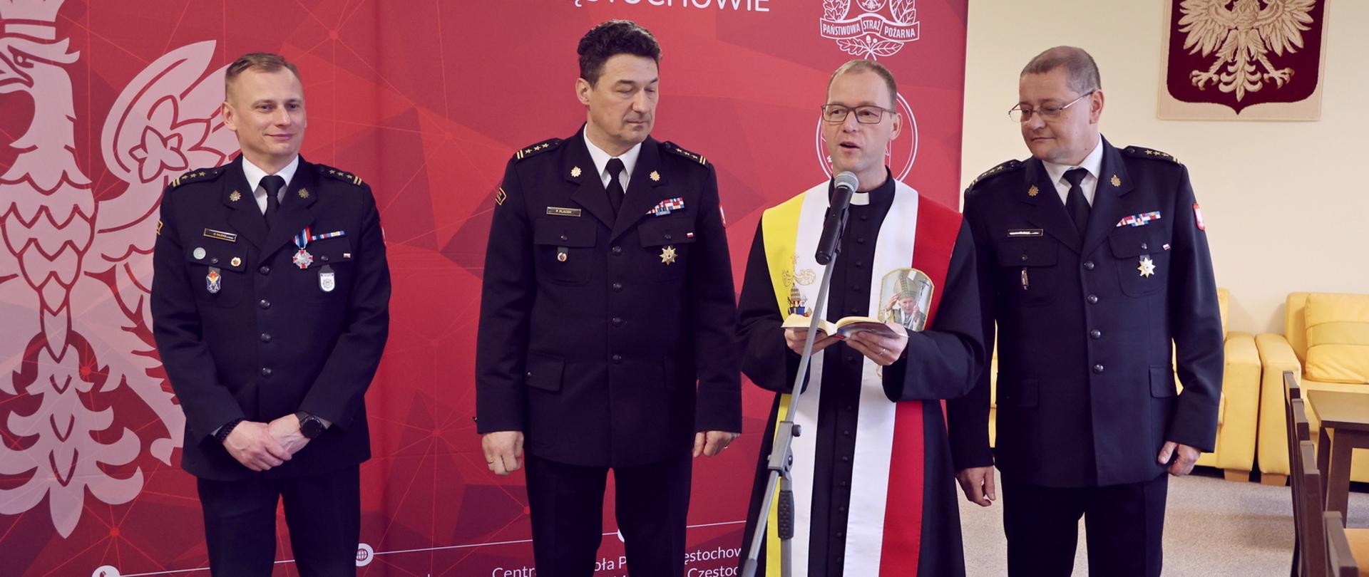 Od lewej stojący Zastępca Komendanta Centralnej Szkoły PSP, Komendant Centralnej Szkoły PSP, ksiądz mówiący do mikrofonu trzymając w dłoni modlitewnik oraz kolejny Zastępca Komendanta Centralnej Szkoły PSP, w tle po prawej godło wiszące na ścianie