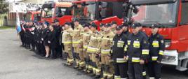 Na zdjęciu widoczni strażacy z Komendy Miejskiej Państwowej Straży Pożarnej w Tarnowie podczas zbiórki na tle samochodów pożarniczych. Strażacy ustawieni w kolejności: poczet sztandarowy, poczet flagowy, funkcjonariusze i pracownicy cywilni oraz strażacy z jednostki ratowniczo-gaśniczej w ubraniach bojowych.