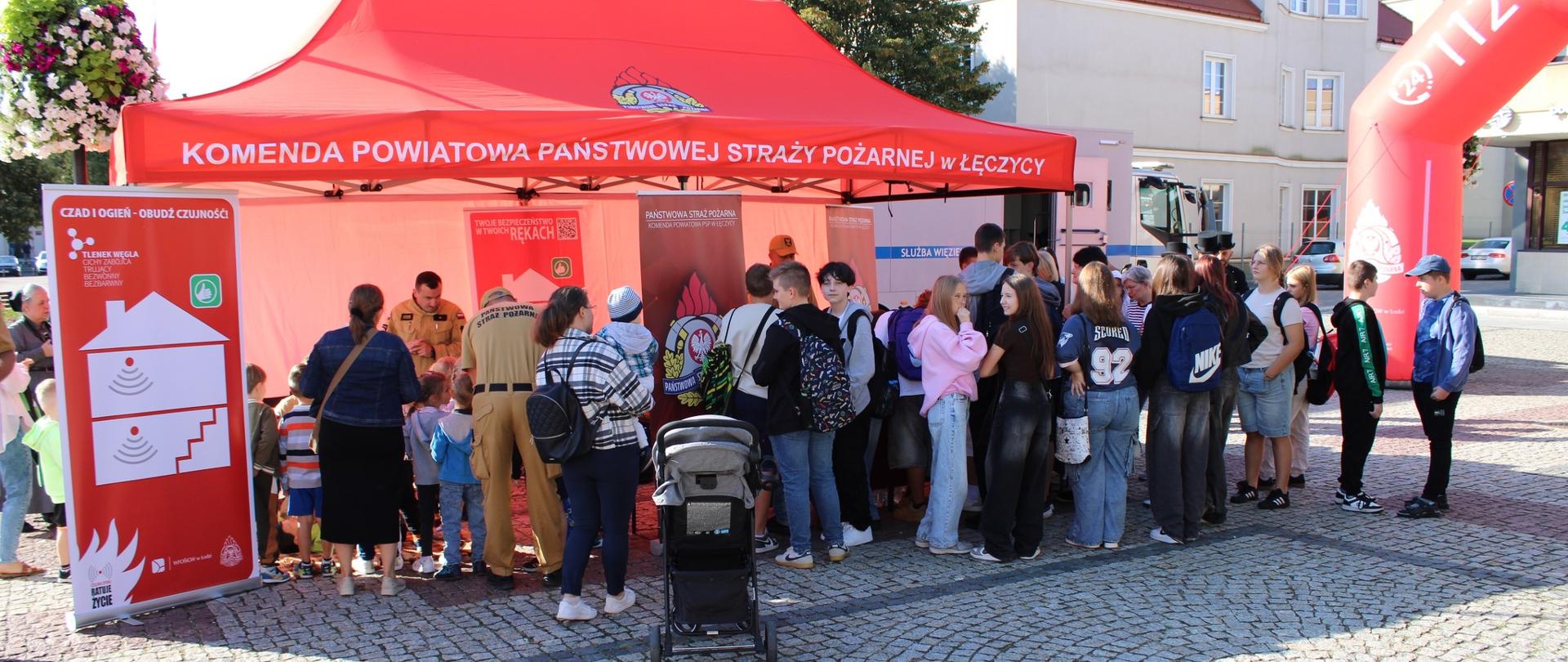 Na zdjęciu widać wydarzenie plenerowe. Na środku ustawiony jest czerwony namiot straży pożarnej, pod którym stoją strażacy w mundurach koloru piaskowego. Przed namiotem widoczna jest spora grupa osób - dzieci i dorośli. Po lewej stronie znajduje się roll-up informacyjny o czujnikach tlenku węgla i zagrożeniach związanych z pożarami, a z prawej strony widoczny jest fragment dużej czerwonej bramy pneumatycznej z numerem alarmowym 112. W tle można dostrzec budynki miejskie oraz zaparkowany pojazd straży pożarnej.