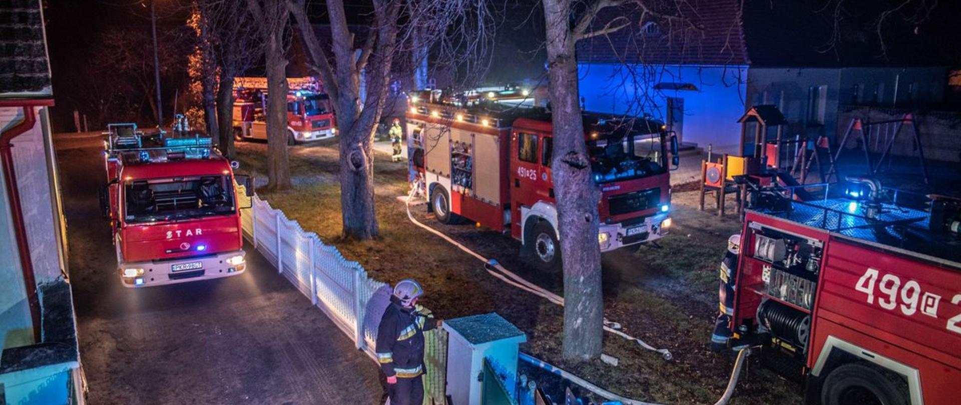 Zdjęcie przedstawia cztery pojazdy pożarnicze stojące na terenie przed posesją, z pojazdów wyprowadzone są węże tłoczne.