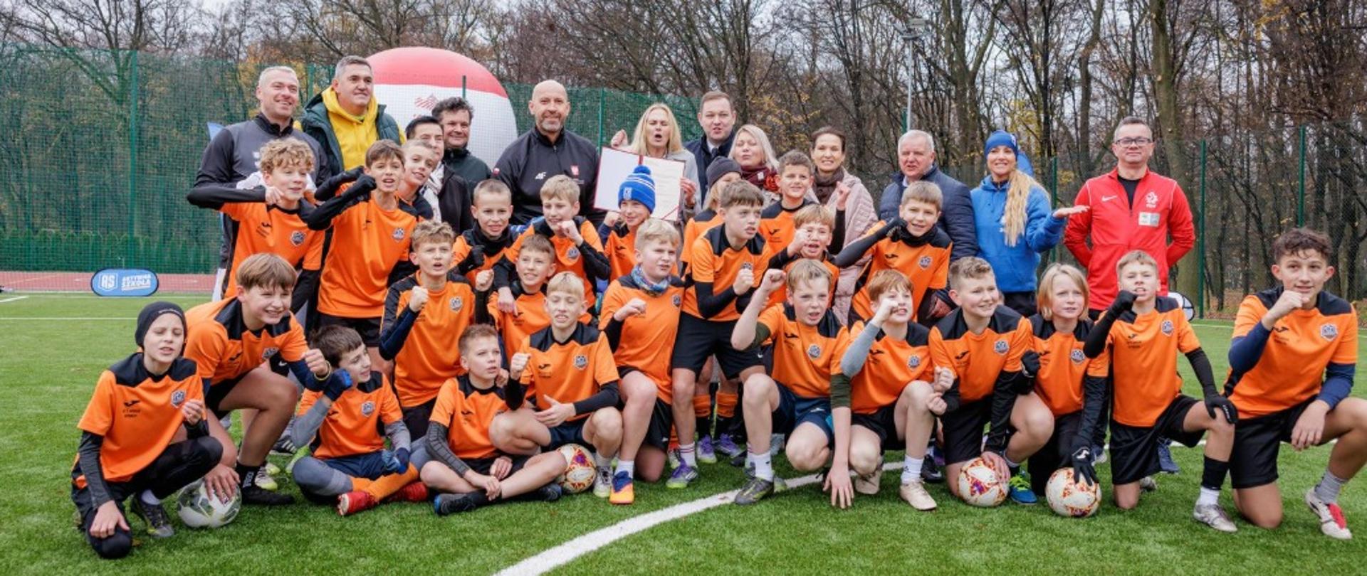 Zdjęcie zbiorowe przedstawiające ministra sportu i turystyki Jakuba Rutnickiego z dziećmi na boisku piłkarskim (Orliku).