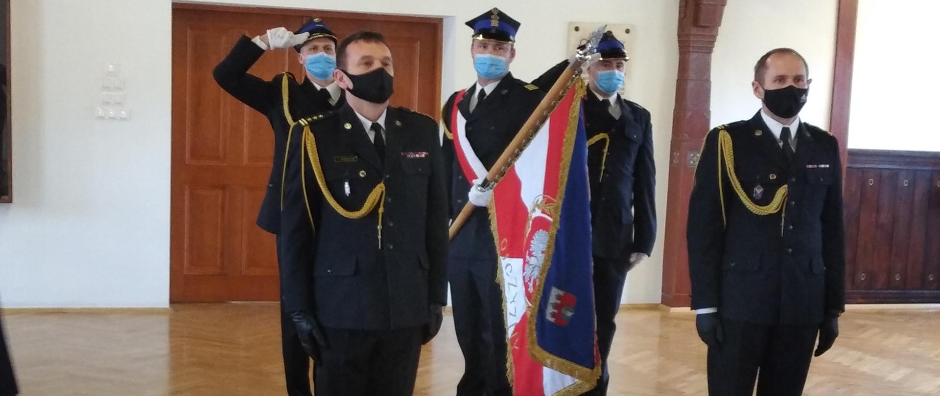 Na pierwszym planie od lewej strony: st. bryg. Jacek Podwolski- zdający obowiązki Zastępcy Komendanta Miejskiego PSP w Bydgoszczy, mł. bryg. Tomasz Czajka- przejmujący obowiązki Zastępcy Komendanta Miejskiego PSP w Bydgoszczy, obydwoje w mundurach wyjściowych i czarnych maseczkach na twarzy. W tle poczet sztandarowy KM PSP w Bydgoszczy, poczet składa się z 3 funkcjonariuszy PSP ubranych w mundury wyjściowe, środkowa osoba z pocztu pochyla sztandar w kierunku przejmującego i zdającego obowiązki. Całość odbywa się w pomieszczeniu świetlicy JRG1 KM PSP w Bydgoszczy.