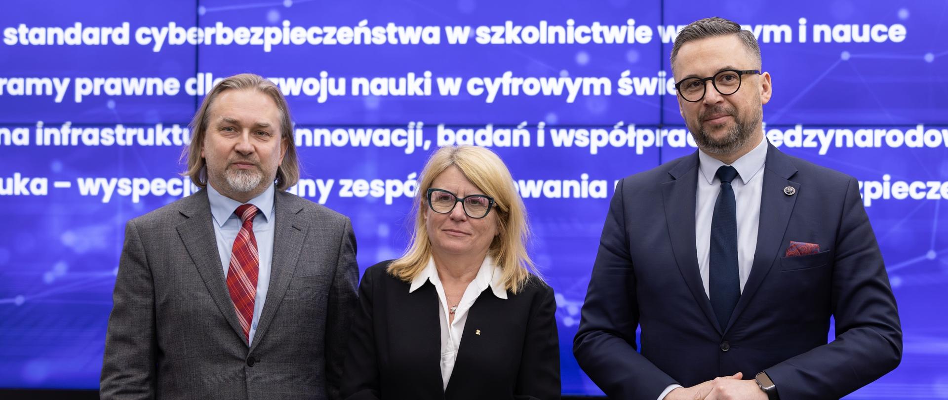 Na tle wielkiego niebieskiego ekranu stoi mężczyzna w szarym garniturze, kobieta w czarnej marynarce i minister Kulasek w ciemnoszarym garniturze.