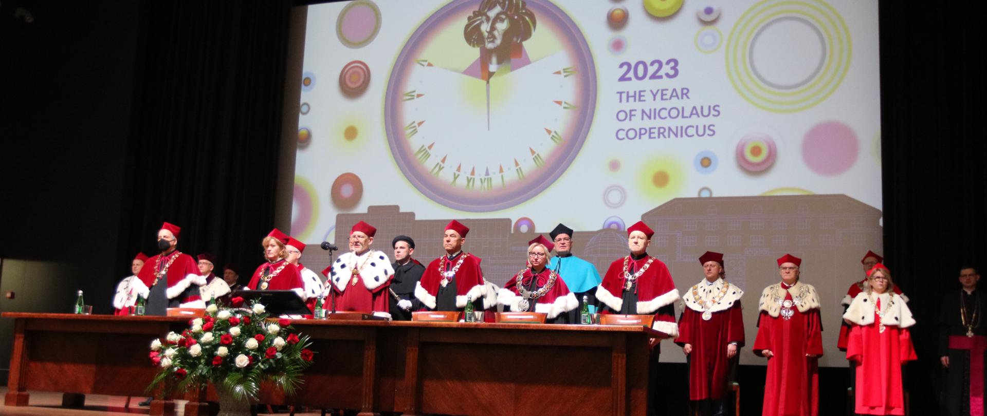 Na podwyższeniu za dużym drewnianym stołem stoi kilkanaście osób w ceremonialnych uniwersyteckich czerwonych strojach, za nimi wielki ekran z napisem 2023 The Year of Nicolaus Copernicus.