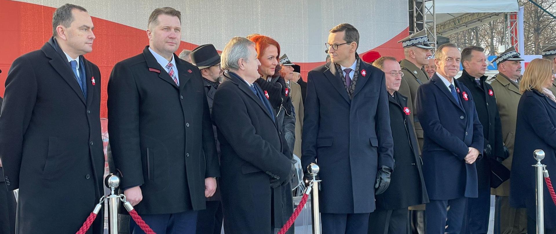 Uroczystości z okazji Narodowego Święta Niepodległości z udziałem ministrem Przemysławem Czarnkiem, minister Przemysław Czarnek stoi na trybunie honorowej podczas uroczystości, z tyłu wyświetlana jest flaga Polski.