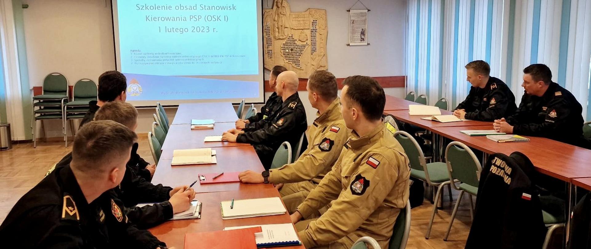 Zdjęcie zrobione w sali wykładowej. Przy stołach konferencyjnych siedzi dziewięciu strażaków uczestniczących w szkoleniu. Za strażakami na ekranie wyświetlony slajd prezentacji z napisem: szkolenie obsad stanowisk kierowania PSP OSK1 1 lutego 2023 roku.