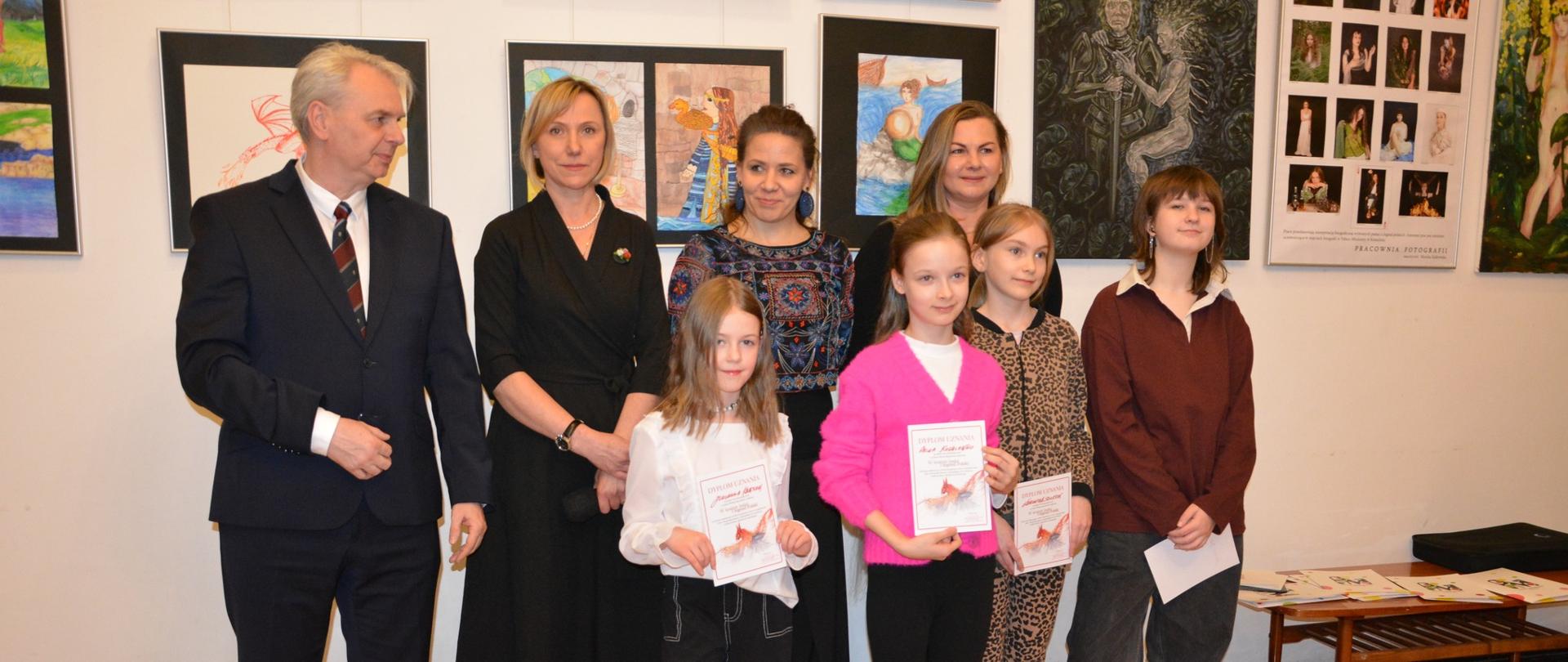Na zdjęciu znajduje się grupa ośmiu osób stojących w dwóch rzędach na tle jasnej ściany galerii. Cztery osoby dorosłe stoją z tyłu, za nimi czwórka dzieci w pierwszym rzędzie. Po lewej stronie widoczny jest mężczyzna w ciemnym garniturze, białej koszuli oraz krawacie w ciemne paski. Obok niego stoi kobieta ubrana w czarną sukienkę do kolan oraz czarne buty na obcasie. Pośrodku znajduje się jeszcze dwóch dorosłych w eleganckich strojach.
Czwórka dzieci na pierwszym planie trzyma w dłoniach dyplomy lub książeczki. Jedno dziecko ma na sobie białą koszulę i ciemne spodnie, kolejne jest ubrane w różowy sweterek oraz czarne spodnie, następne nosi strój w cętki, a ostatnie ma bordową bluzę oraz szerokie, ciemne spodnie i sportowe buty. W tle na ścianie wiszą liczne kolorowe prace plastyczne przedstawiające m.in. postacie, pejzaże i sceny fantastyczne. Po prawej stronie widać niewielki stolik z ułożonymi dokumentami i broszurami. Podłoga w pomieszczeniu jest wyłożona jasnobrązową, błyszczącą wykładziną.