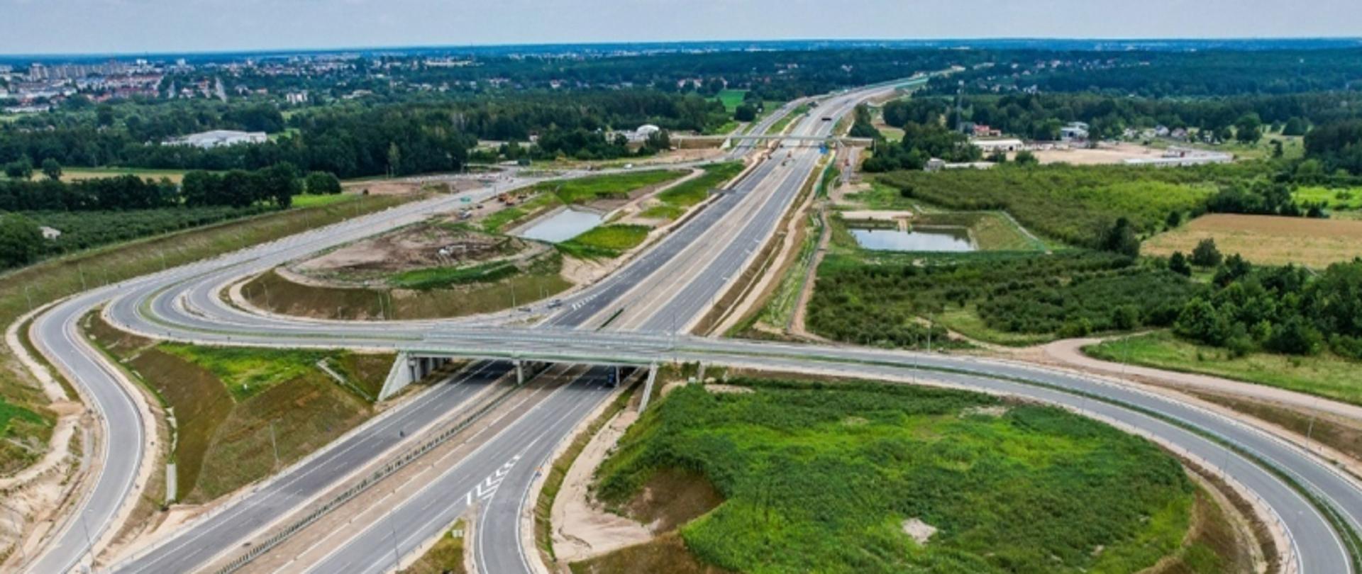 Aerial view węzła autostradowego z licznymi połączeniami drogowymi wśród zielonych pól i zabudowań.