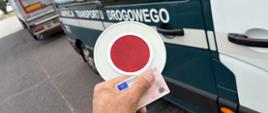 Na pierwszym planie: funkcjonariusz mazowieckiej Inspekcji Transportu Drogowego trzyma w ręku blankiet prawa jazdy kontrolowanego kierowcy i tarczę (lizak) do zatrzymywania pojazdów w ruchu drogowym. W tle: prawy bok oznakowanego furgonu mazowieckiej Inspekcji Transportu Drogowego. Za oznakowanym radiowozem Inspekcji Transportu Drogowego stoi ciągnik siodłowy z podpiętą naczepą.