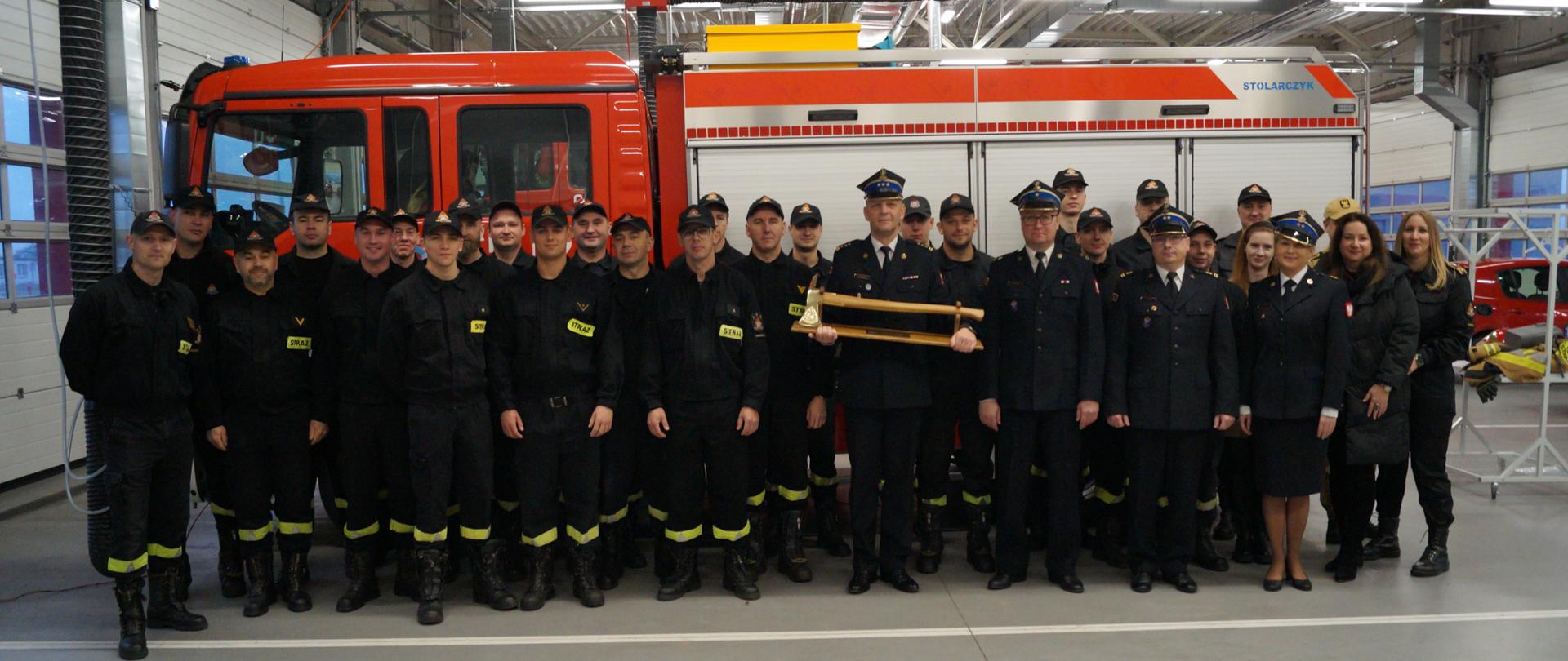 Zdjęcie przedstawia grupę strażaków na tle wozu pożarniczego. 