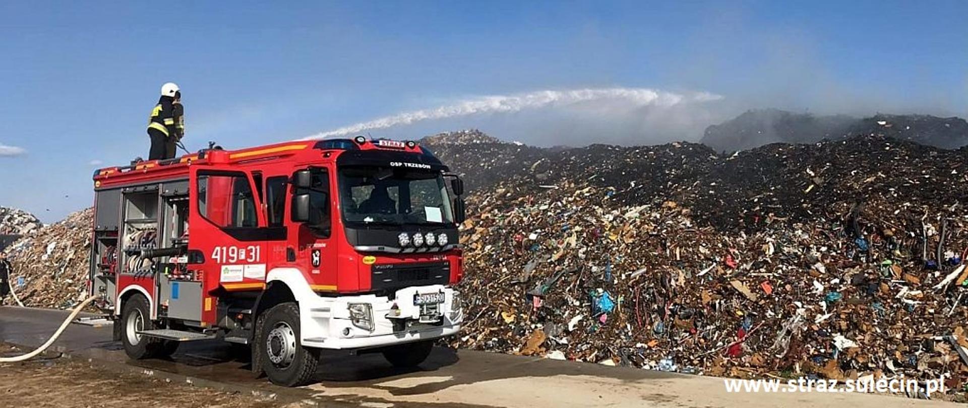 Na zdjęciu znajduje się wóz strażacki na którym znajduje się strażak gaszący stertę śmieci.