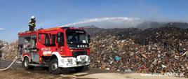 Na zdjęciu znajduje się wóz strażacki na którym znajduje się strażak gaszący stertę śmieci.