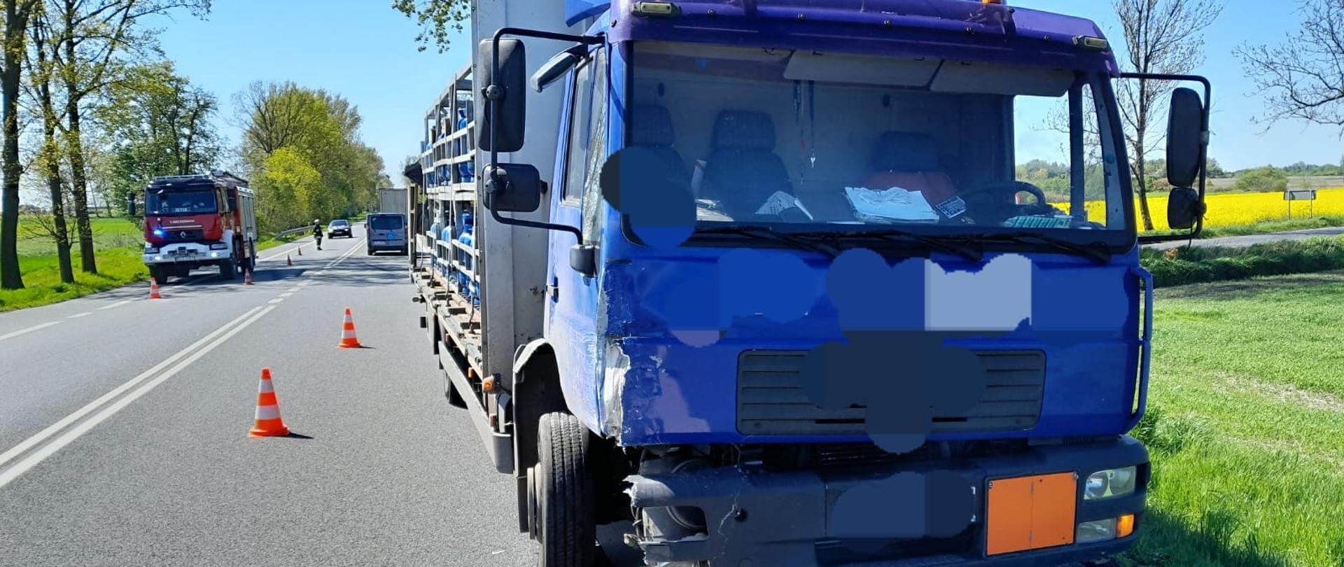 Na zdjęciu widać uszkodzony niebieski samochód dostawczy, który przewozi butle z propan-butanem. Samochód stoi na asfaltowej drodze. Obok rozstawione są biało-pomarańczowe pachołki drogowe. W tle stoi czerwony samochód strażacki. W oddali widać strażaka kierującego ruchem drogowym, samochody oraz drzewa. Jest widno.