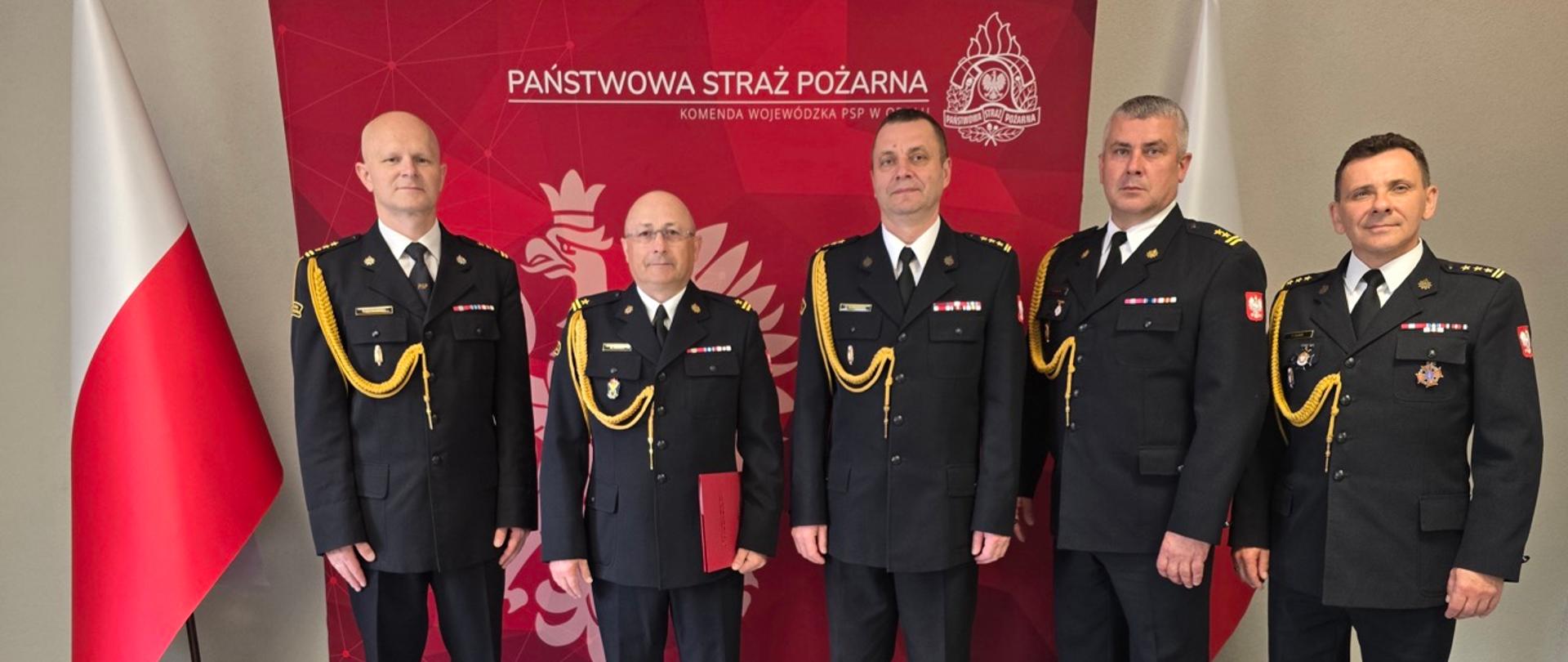 Pięciu wysokich rangą oficerów Państwowej Straży Pożarnej stoi w równym szeregu przed czerwoną ścianką z godłem Polski i napisem „Państwowa Straż Pożarna, Komenda Wojewódzka PSP w Opolu”. Wszyscy mają na sobie galowe mundury z odznaczeniami i złotymi kordami. Wśród nich znajdują się: st. bryg. Arkadiusz Kuśmierski, st. bryg. Piotr Dudek, bryg. Marcin Skalny, st. bryg. Paweł Gotkowski oraz mł. bryg. Zenon Błoch. Po obu stronach stoją polskie flagi. Zdjęcie przedstawia oficjalną, reprezentacyjną fotografię uczestników uroczystości powołania nowego zastępcy komendanta powiatowego PSP w Nysie.