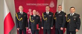 Pięciu wysokich rangą oficerów Państwowej Straży Pożarnej stoi w równym szeregu przed czerwoną ścianką z godłem Polski i napisem „Państwowa Straż Pożarna, Komenda Wojewódzka PSP w Opolu”. Wszyscy mają na sobie galowe mundury z odznaczeniami i złotymi kordami. Wśród nich znajdują się: st. bryg. Arkadiusz Kuśmierski, st. bryg. Piotr Dudek, bryg. Marcin Skalny, st. bryg. Paweł Gotkowski oraz mł. bryg. Zenon Błoch. Po obu stronach stoją polskie flagi. Zdjęcie przedstawia oficjalną, reprezentacyjną fotografię uczestników uroczystości powołania nowego zastępcy komendanta powiatowego PSP w Nysie.