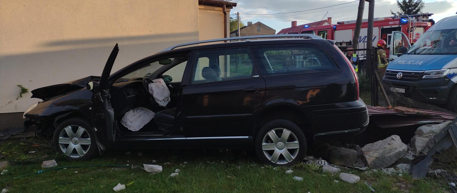 Na pierwszym planie samochód osobowy który uderzył w ścianę budynku, uszkodzona studnia. Za pojazdem w tle znajdują się strażacy i policjanci , w tle radiowóz policyjny , i 2 wozy strażacki. 