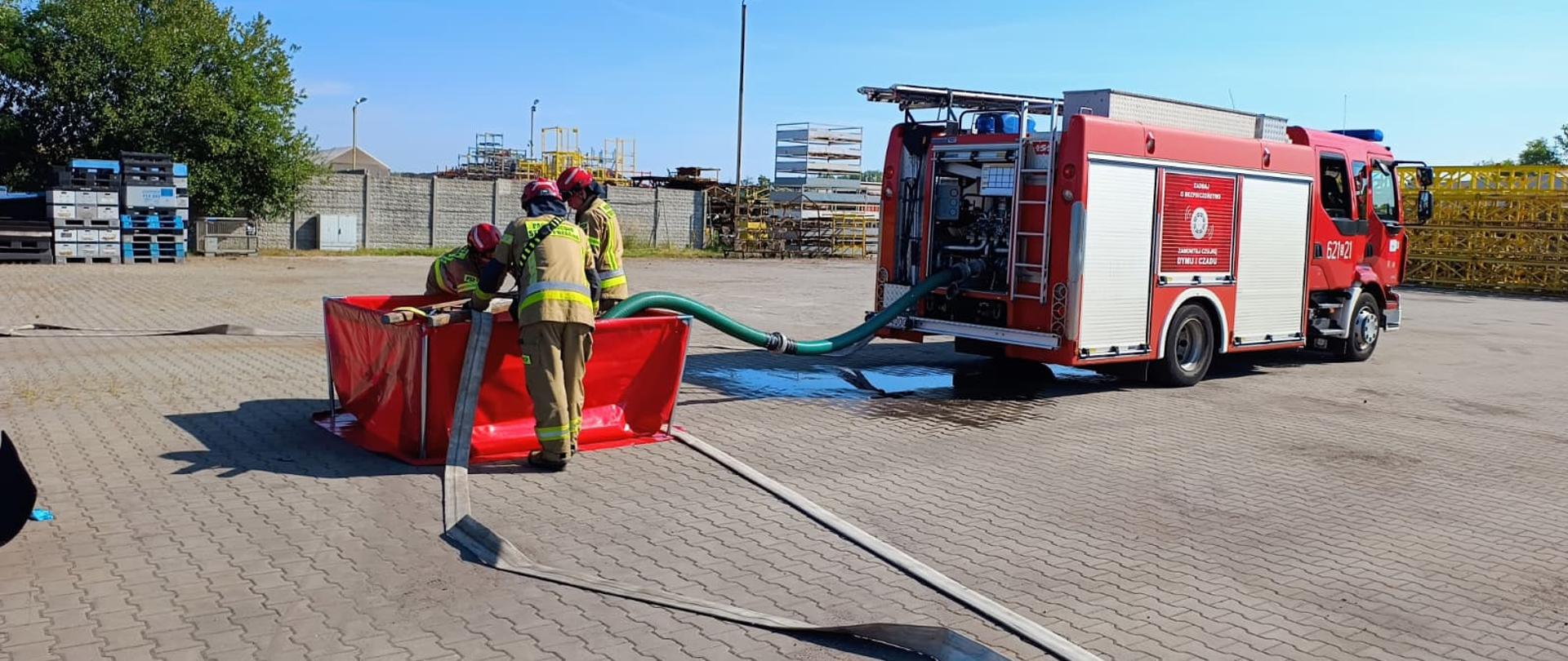 Na pierwszym planie, po lewej stronie, widać trzech strażaków w pełnym umundurowaniu ochronnym – mają na sobie żółte ubrania, hełmy i spodnie. Dwóch z nich jest pochylonych nad czerwonym, składanym zbiornikiem na wodę, do którego jest podłączony gruby, zielony wąż. Wokół widać porozrzucane inne węże strażackie. Trwają ćwiczenia. Woda rozlewa się po betonowym placu.
W tle stoi duży, czerwony wóz strażacki z numerem 621-21 na drzwiach. Widać, że jest on źródłem wody dla zbiornika. Scena rozgrywa się na dużym, utwardzonym placu, otoczonym z jednej strony betonowym murem, a z drugiej różnymi konstrukcjami i magazynami. Panuje słoneczna pogoda, a niebo jest bezchmurne.
