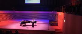 Uczeń gra na puzonie stojąc przed fortepianem, akompaniatorka gra na fortepianie na scenie sali koncertowej PSM
