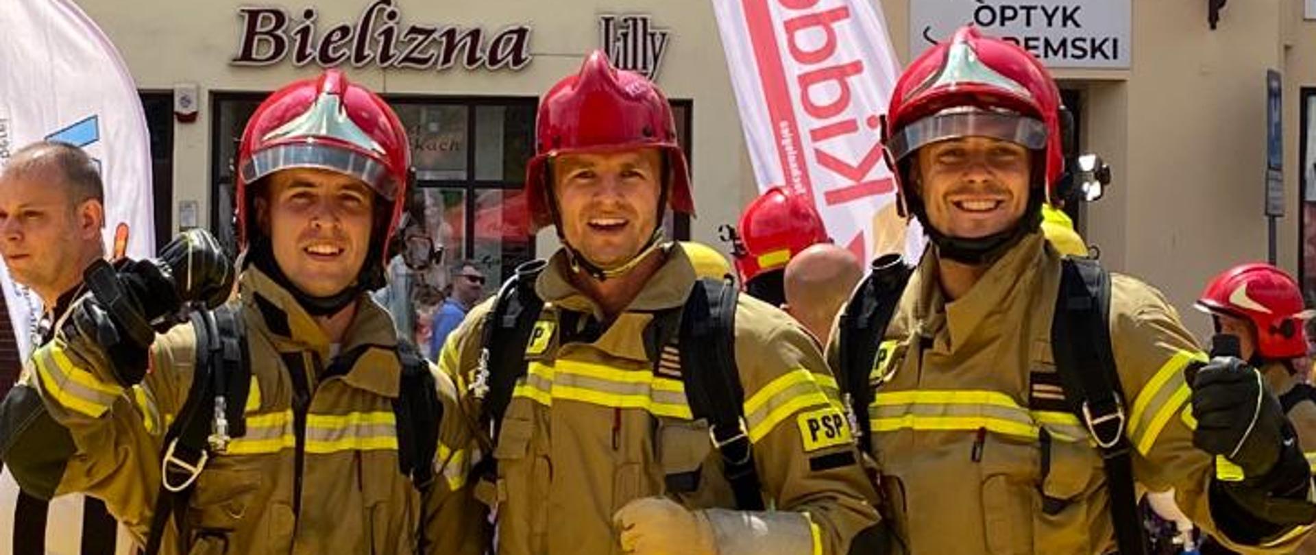 Trzech strażaków pozuje do zdjęcia. W tle widoczni widzowie oraz budynki.
