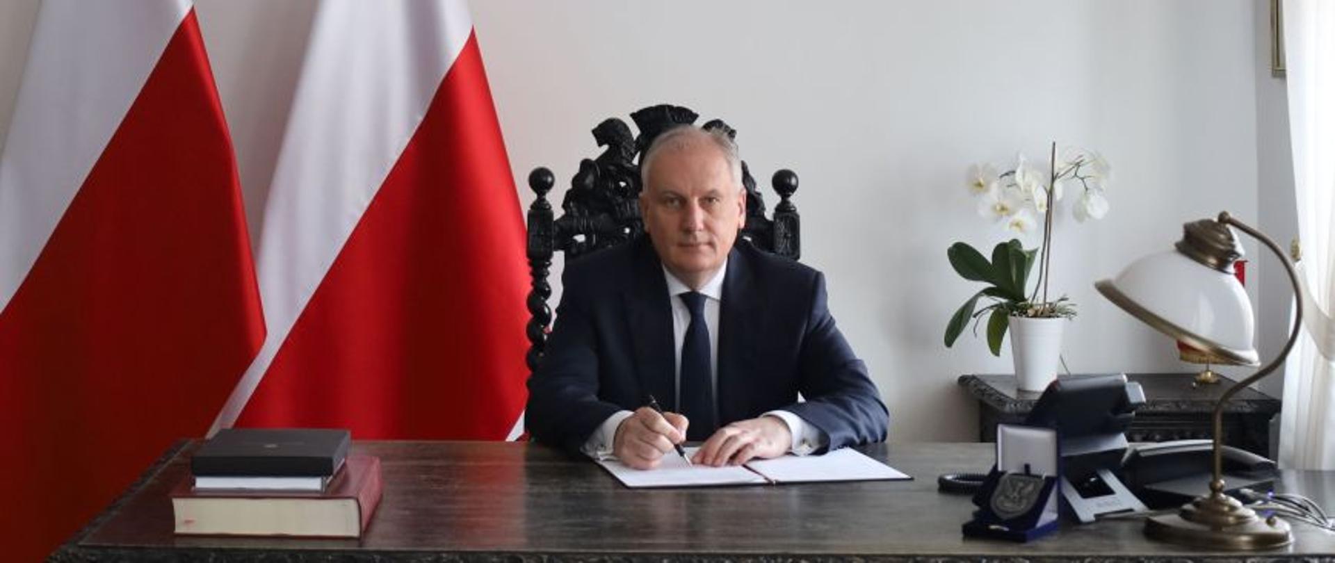 Mężczyzna siedzi za biurkiem podpisując dokument za nim są dwie flagi Polski na ścianie wisi godło Polski.