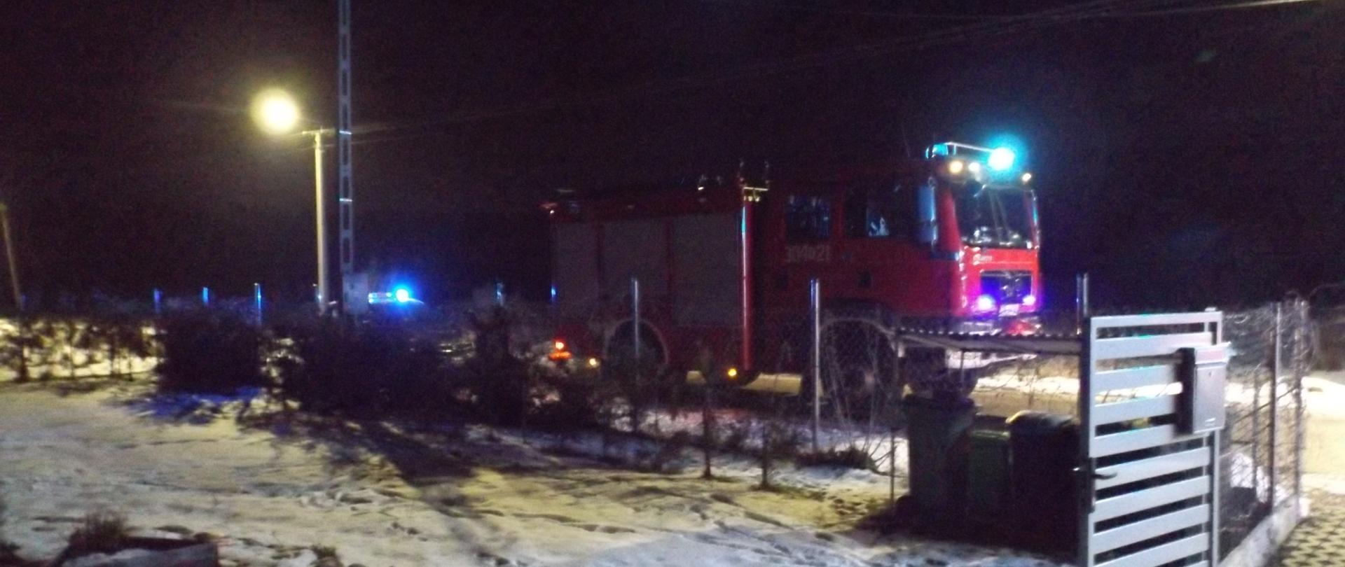 Na zdjęciu widać samochód strażacki znajdujący się przed ogrodzeniem posesji, na której doszło do pożaru.