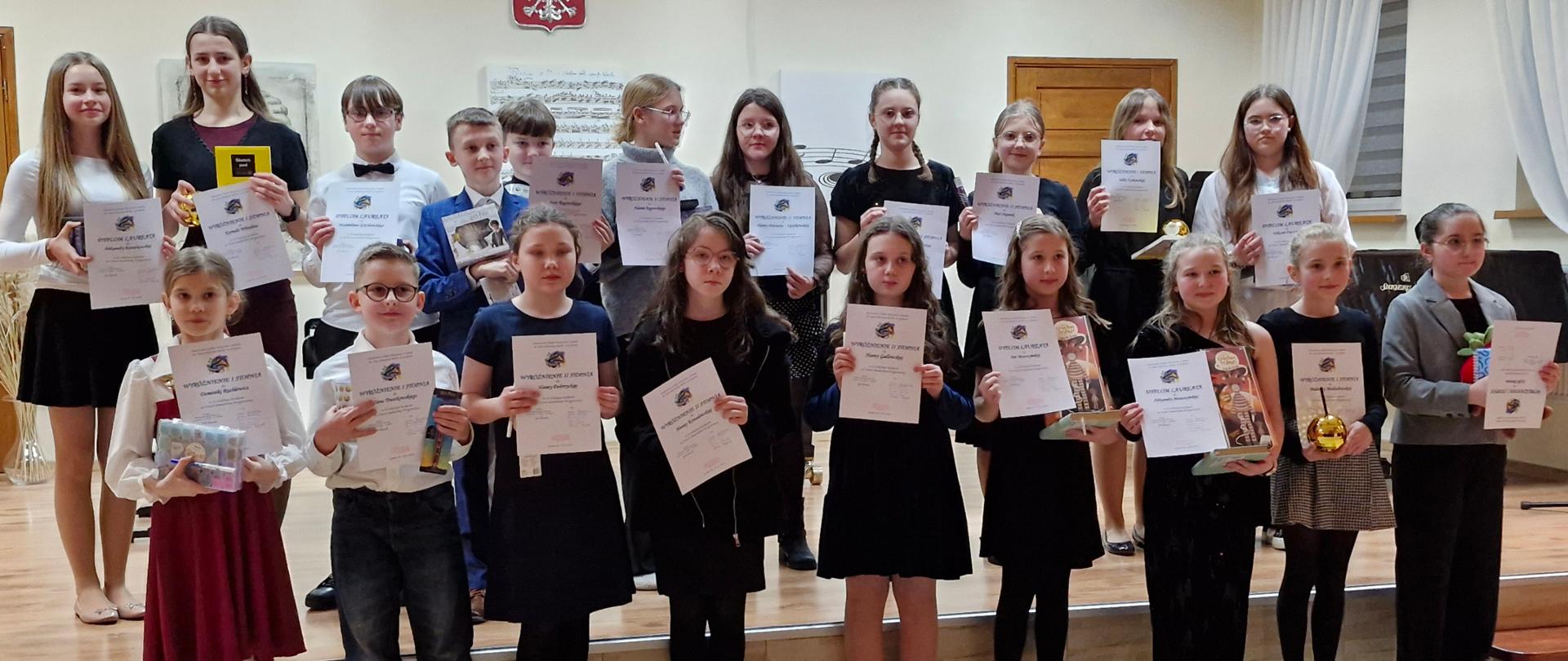 Zdjęcie przedstawia laureatów Konkursu w II Kategorii. Dzieci stoją w dwóch rzędach , trzymają w rękach dyplomy. W tle Godło, drzwi, okno z firankami.