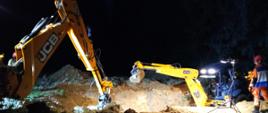 Na zdjęciu w nocy widoczne dwie koparki koloru żółtego prowadzące czynności zmierzające do odsunięcia od zbocza wykopu urobku obciążającego w trakcie działań ratowniczych.