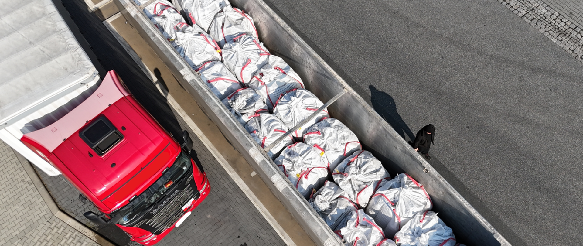 Samochód ciężarowy na parkingu, obok niego odsłonięta naczepa, w której znajduje się duża ilość dużych worków. 