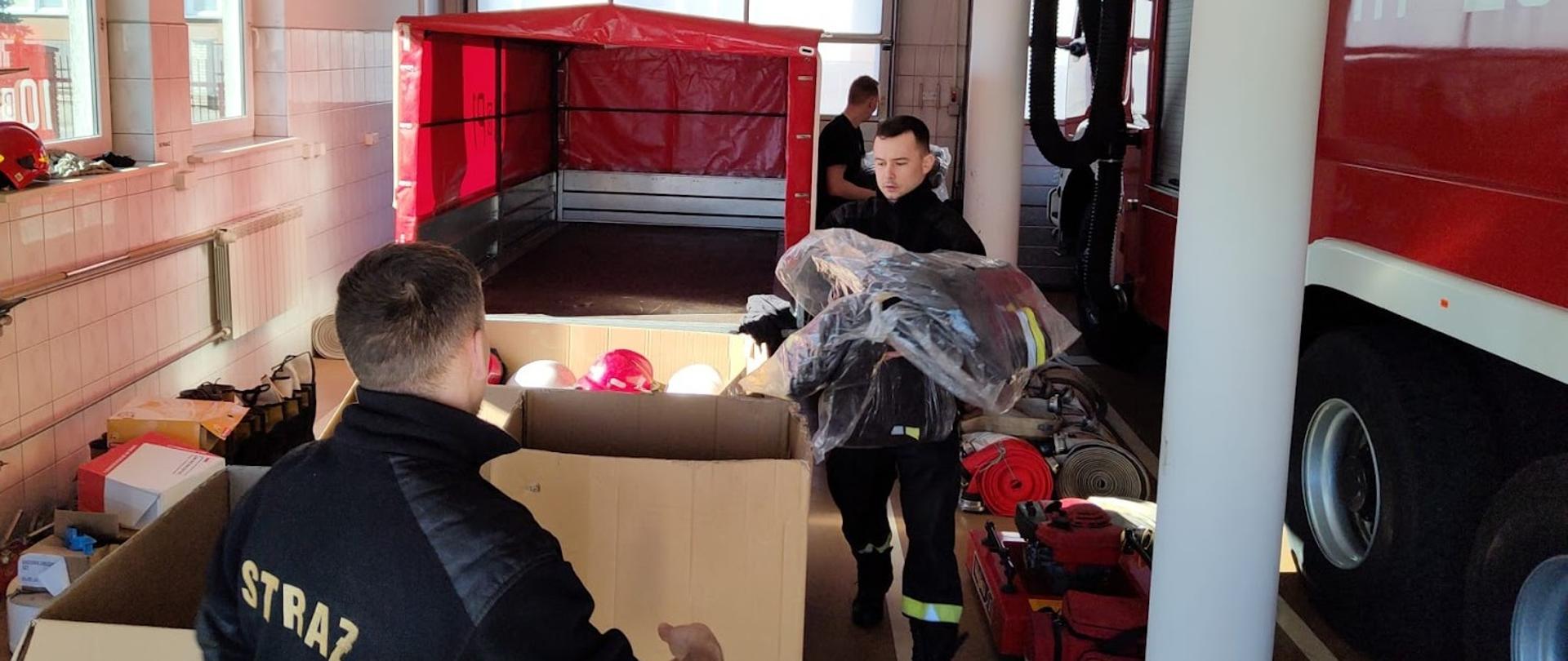 zdjęcie przedstawia trzech strażaków pakujących do kartonu czarną odzież specjalną NOMEX w tle widać wóz ratowniczo-gaśniczy oraz przyczepkę z czerwoną plandeką 
