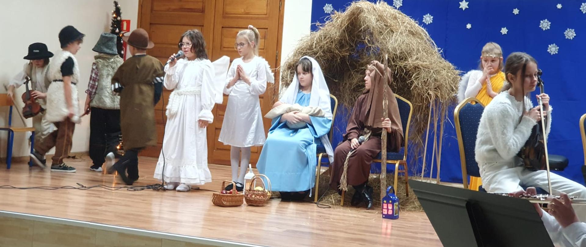 Na scenie uczniowie przebrani za Maryję i Józefa siedzą na krzesłach a w tle szopka. 