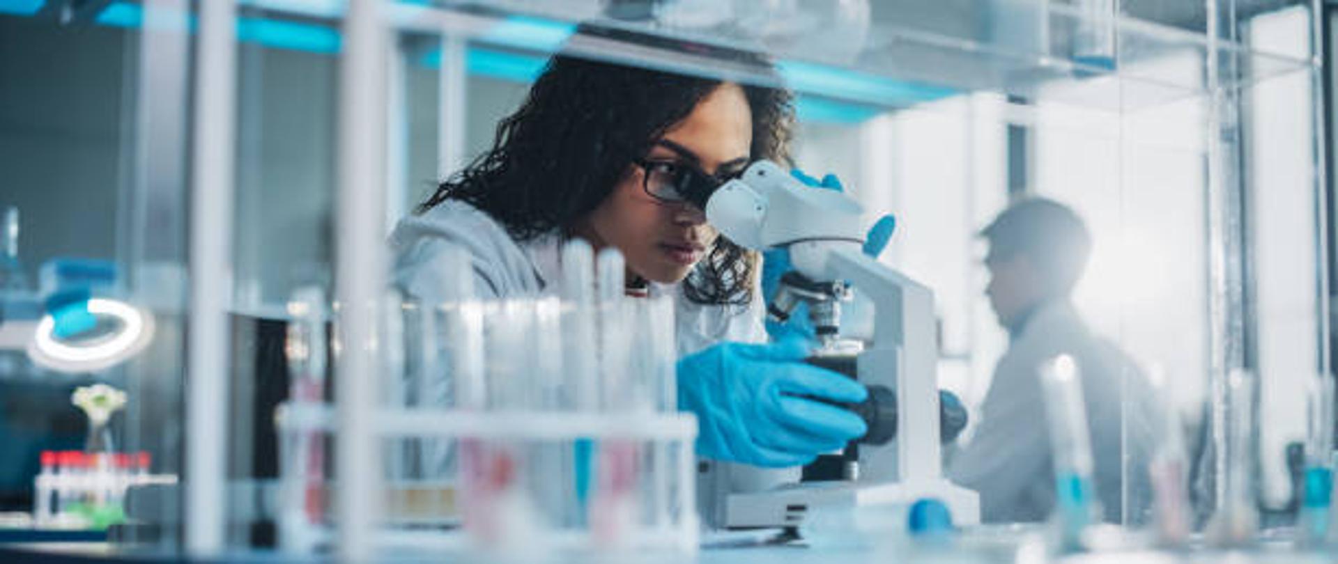 Laborantka patrzy w mikroskop i analizuje próbkę. Na stoliku stoją probówki i wyposażenie laboratorium.
