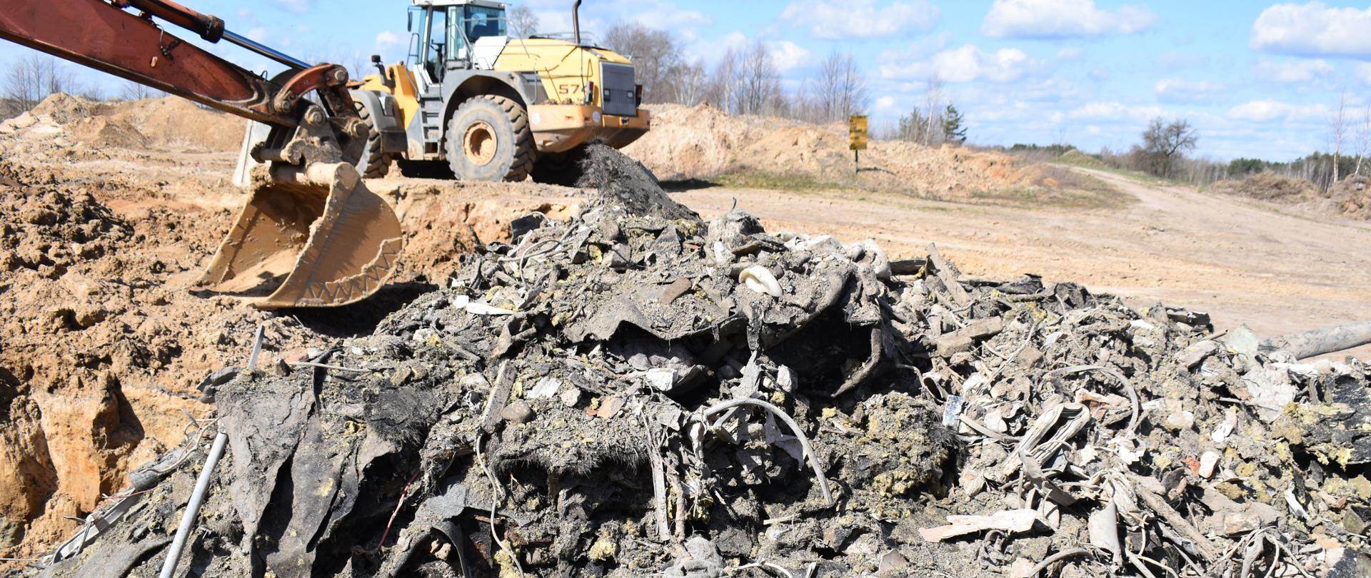 Zmieszane odpady budowlane zalegają na nieutwardzonym, piaszczystym podłożu. W tle łyżka koparki oraz spychacz.