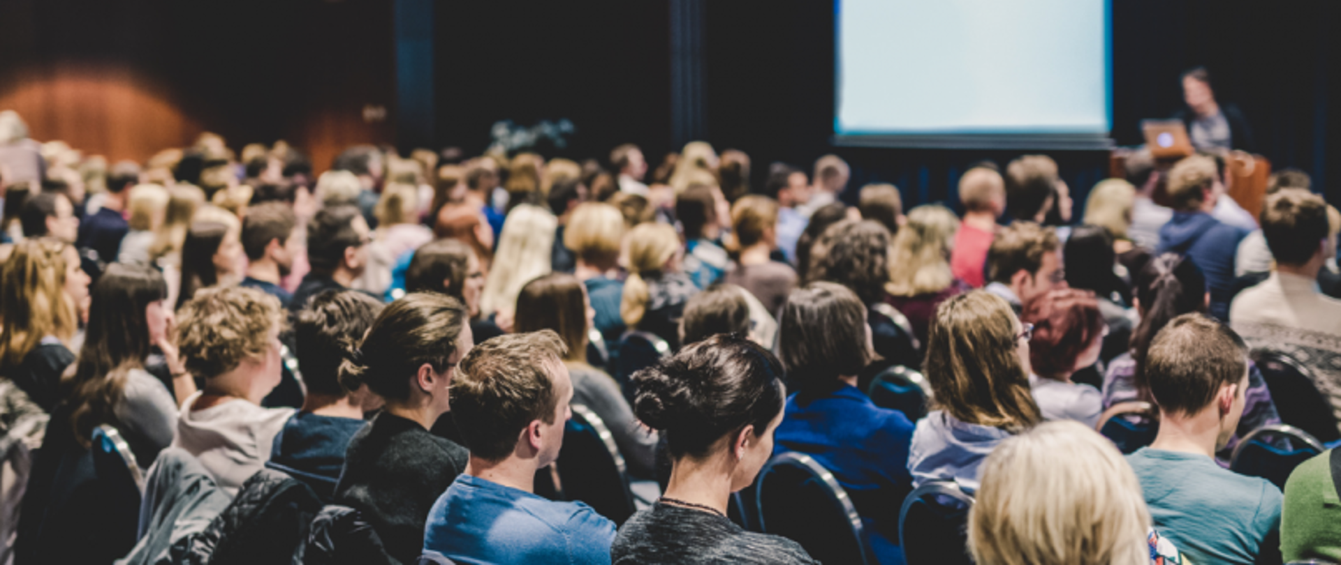 Publiczność siedząca w sali konferencyjnej, w tle prelegent i ekran prezentacyjny, lekko rozmyte.