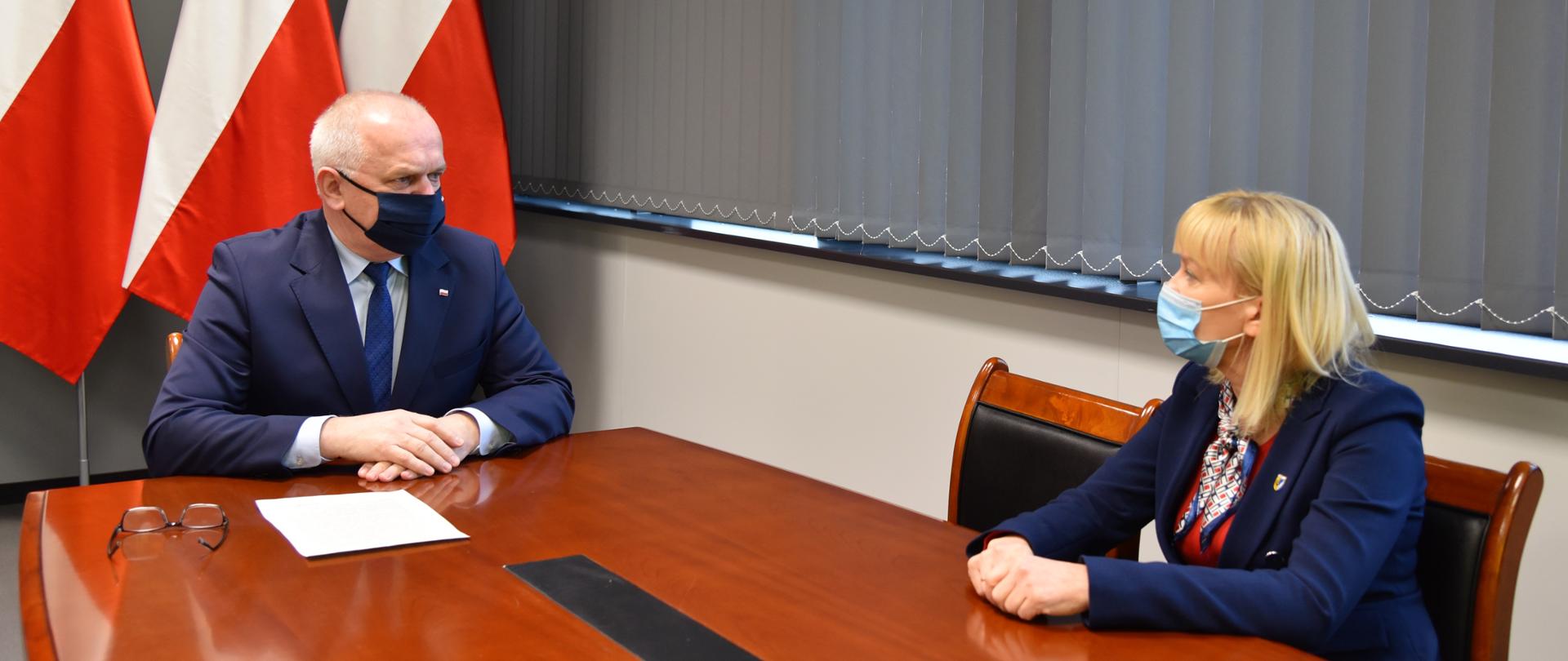 Wojewoda Władysław Dajczak rozmawia ze starostą powiatu nowosolskiego Iwoną Brzozowską. Siedzą przy stole, w tle flagi