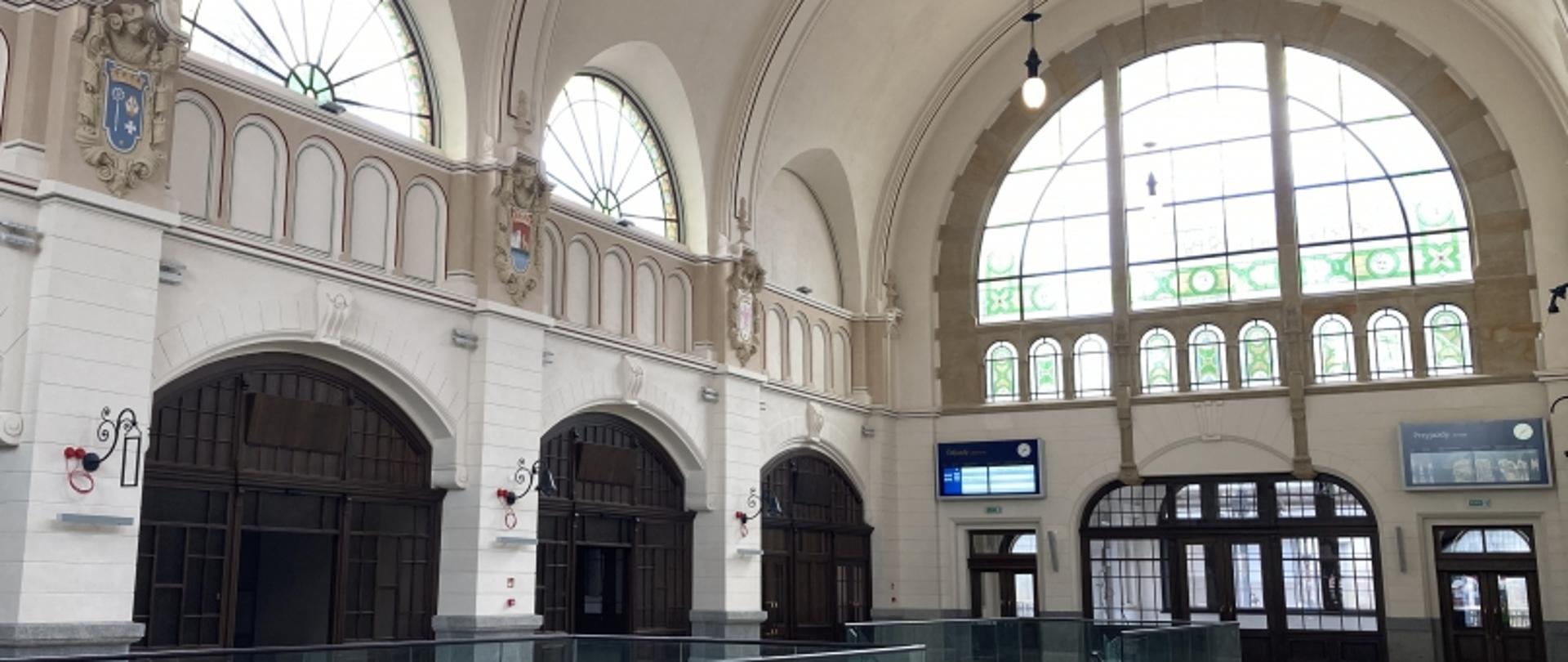 Zabytkowe wnętrze z łukowymi sklepieniami, dużymi oknami, drewnianymi drzwiami i ekranami informacyjnymi; naturalne światło rozświetla przestrzeń.