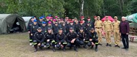 Pamiątkowe zdjęcie uczestników obozu Młodzieżowej Drużyny Pożarniczej Ochotniczej Straży Pożarnej w Łebuni z Zastępcą Pomorskiego Komendanta Wojewódzkiego Państwowej Straży Pożarnej, Komendantem Powiatowym Państwowej Straży Pożarnej w Lęborku, Dowódcą Jednostki Ratowniczo - Gaśniczej oraz Wójtem Gminy Cewice. W tle widoczny teren obozowiska w lesie oraz namioty obozowe.