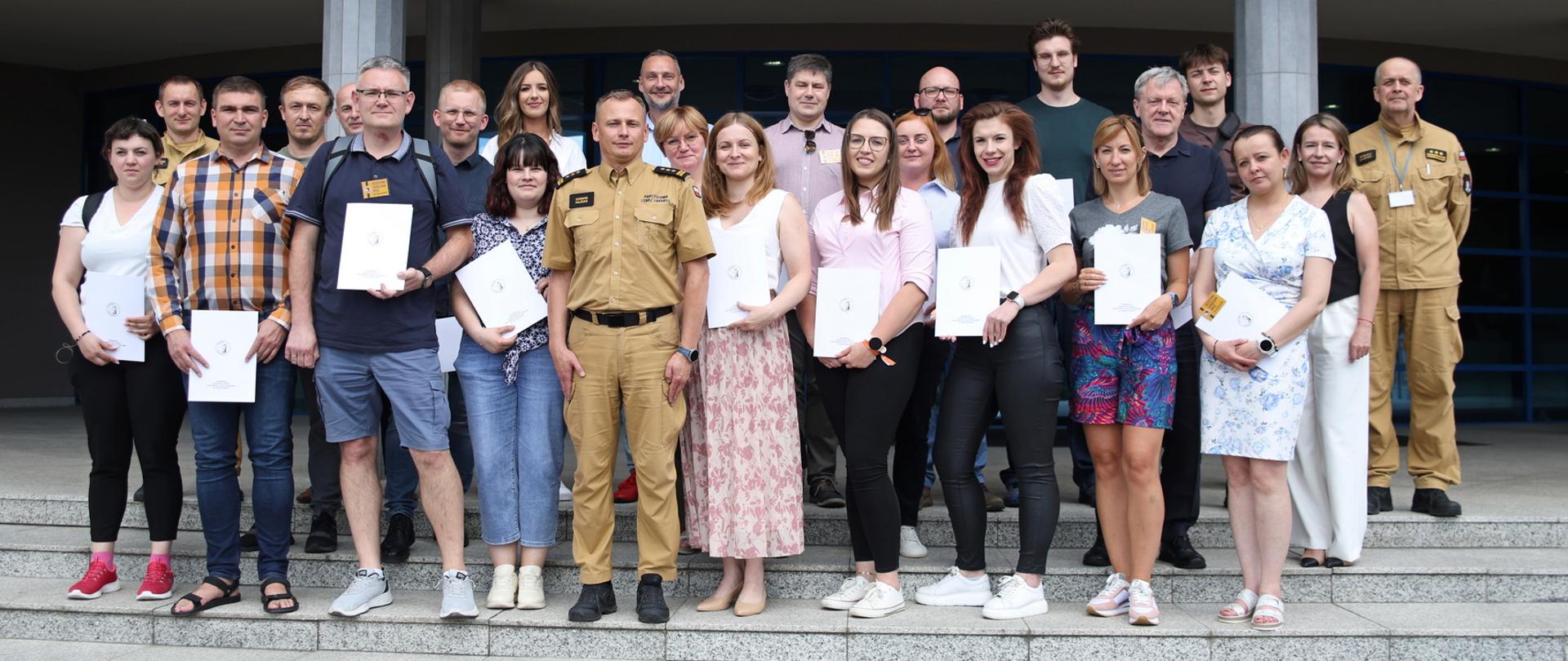 Pamiątkowe zdjęcie grupowe uczestników szkolenia inspektorów ochrony przeciwpożarowej (21 osób) w towarzystwie Zastępcy Komendanta Centralnej Szkoły PSP, kobiety oraz dwóch funkcjonariuszy CS PSP