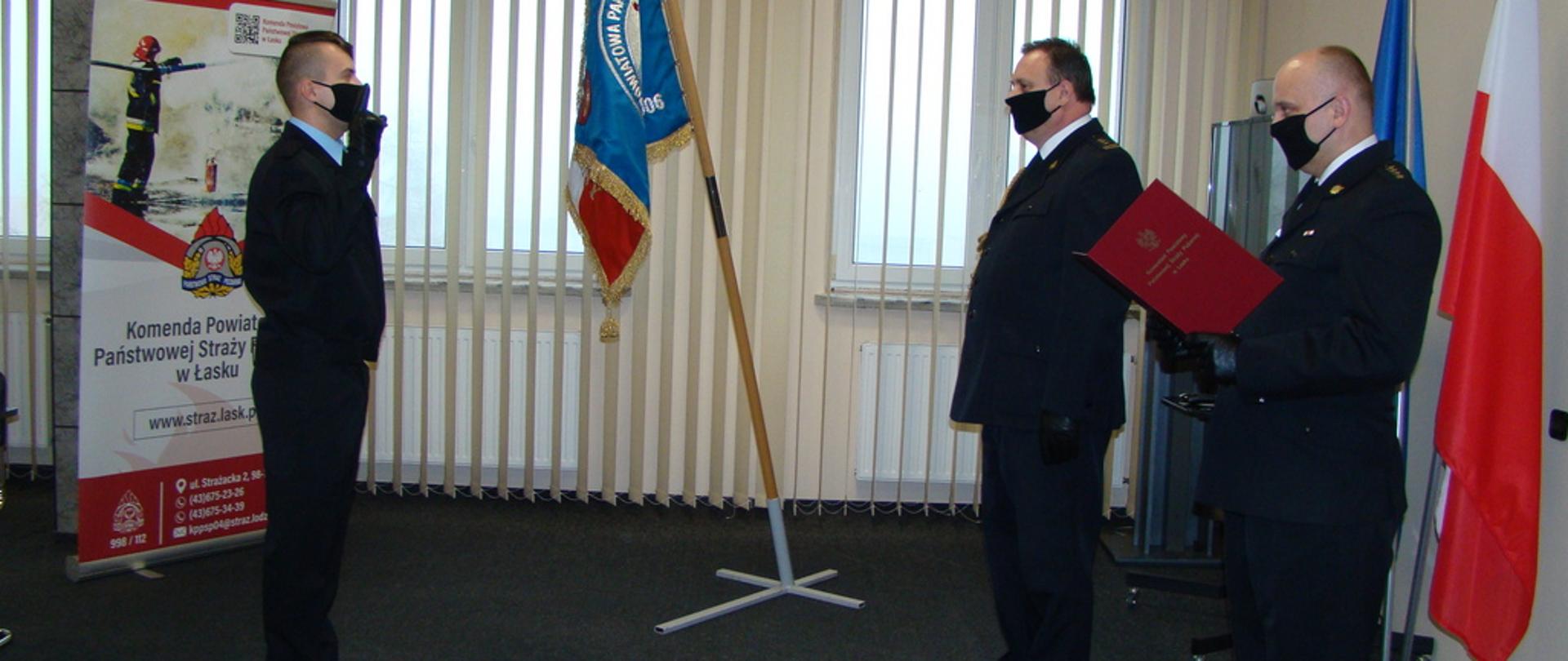 Zdjęcie przedstawia uroczystość ślubowania nowego strażaka w Komendzie Powiatowej Państwowej Straży Pożarnej w Łasku. Na zdjęciu widoczni są Komendant Powiatowy PSP , Zastępca Dowódcy Jednostki Ratowniczo Gaśniczejj oraz strażak wypowiadający słowa ślubowania. 